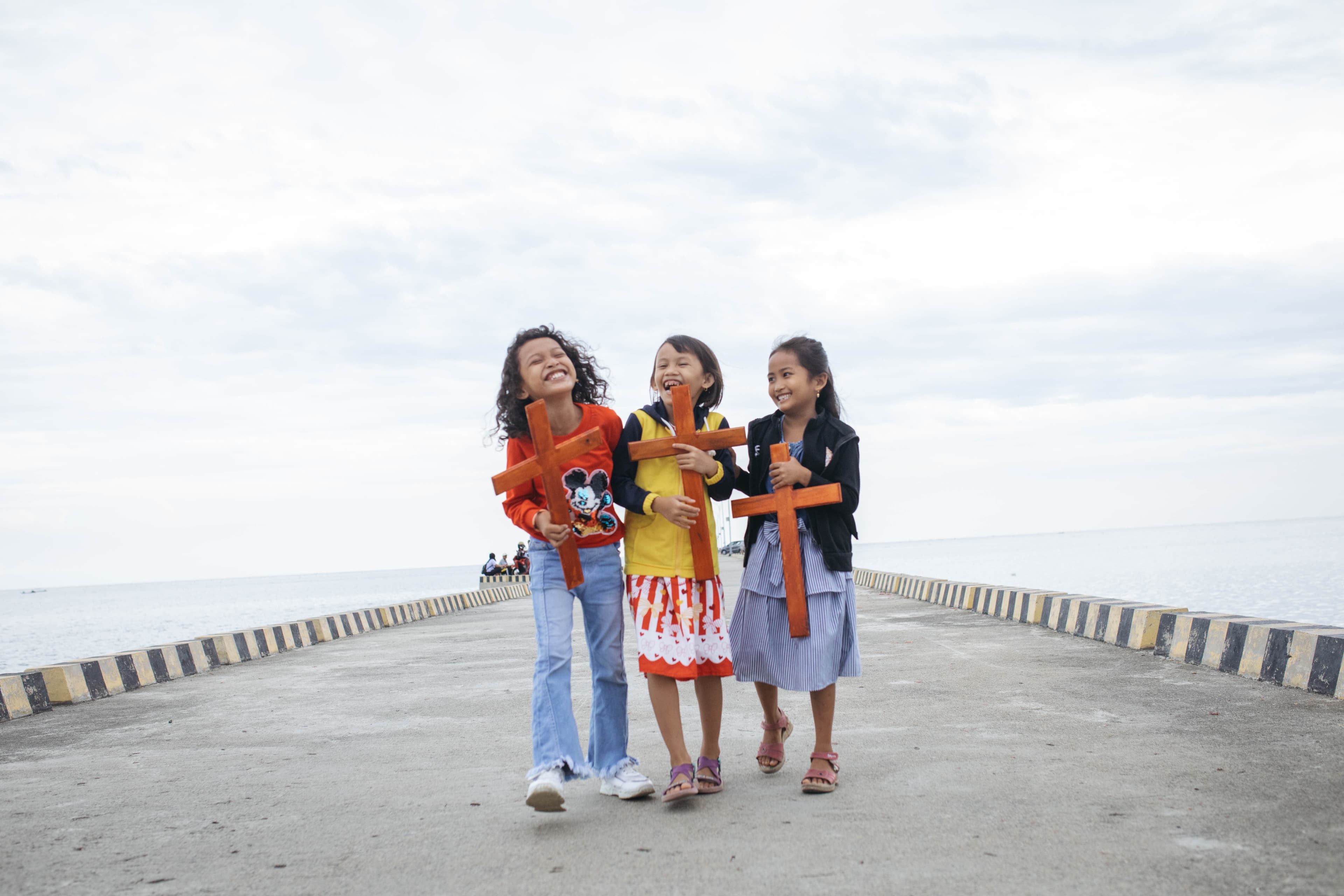 Three little girls walk on an ocean boardwalk toward the camera holding wooden crosses and smiling.