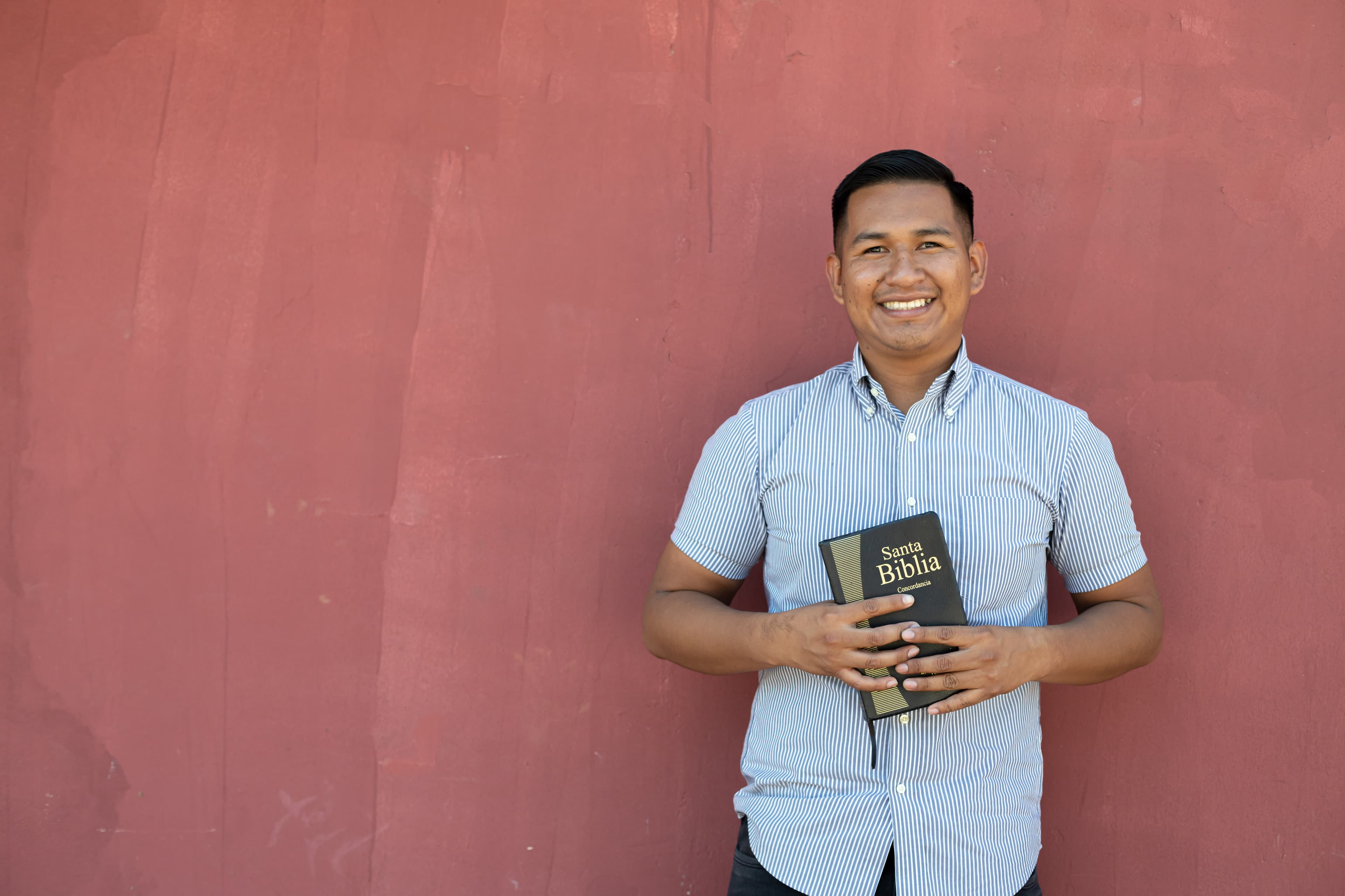 An alumnus of the Compassion program is wearing a gray and white shirt and smiling and holding a Bible. He is standing in front of a pink wall.
