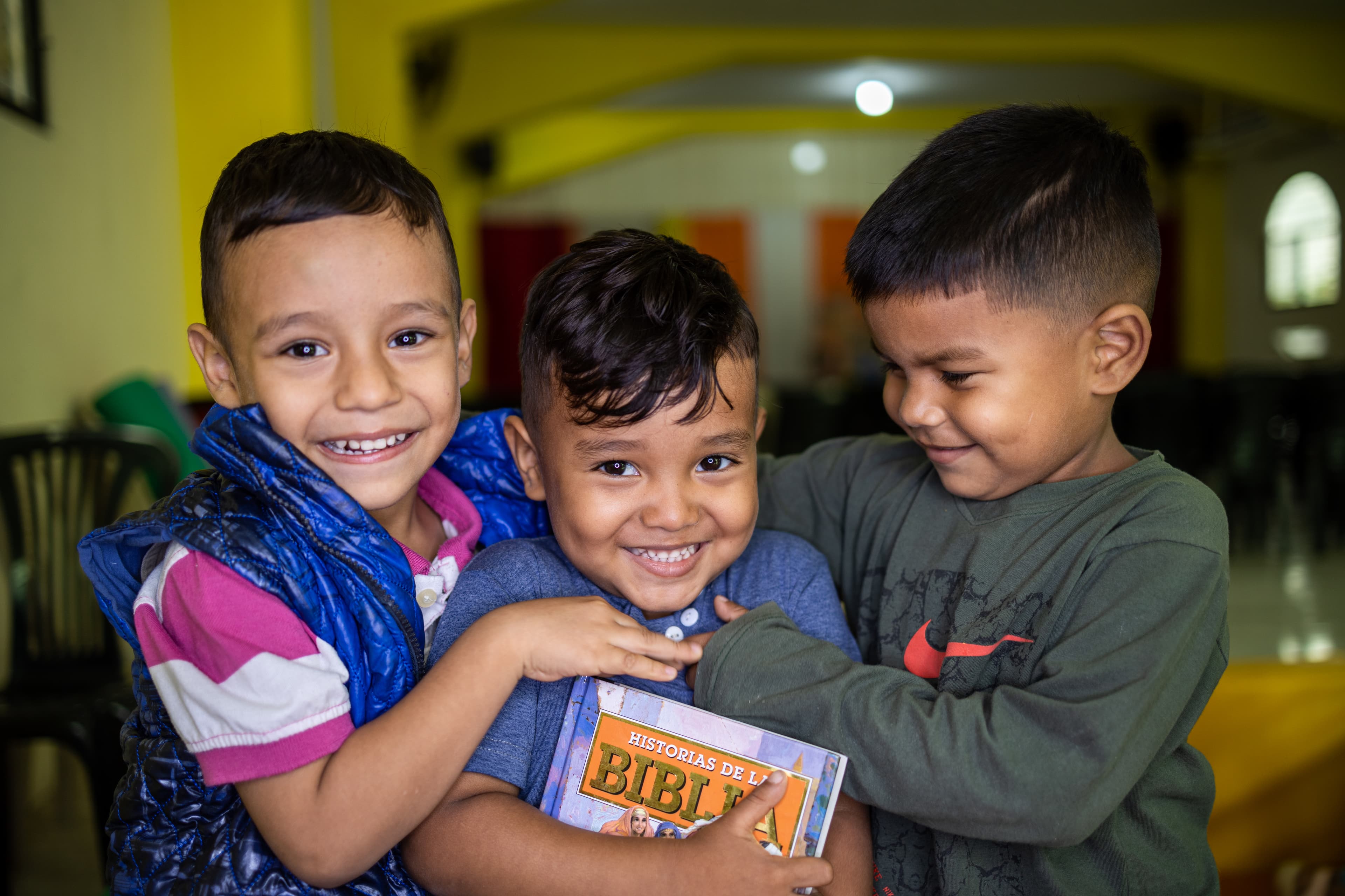 Three young boys hug each other and smile while one holds a Bible.