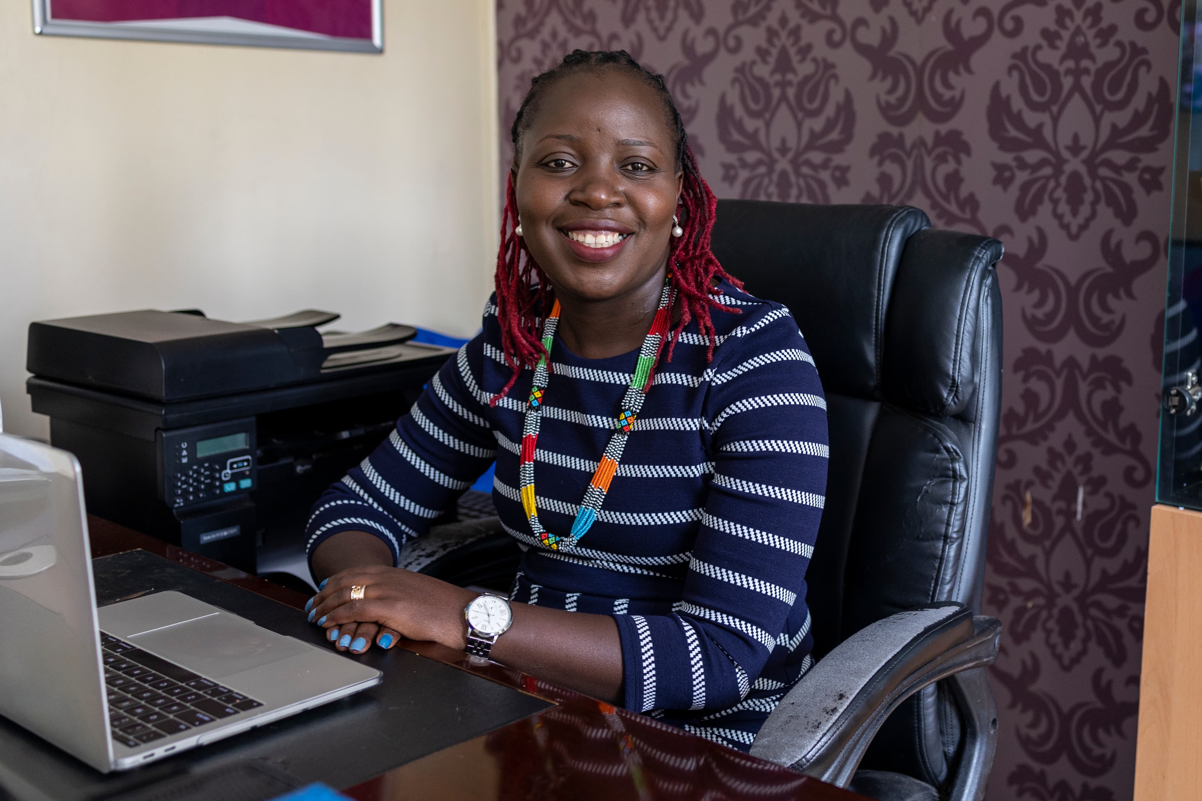 A young woman sits in front of a laptop in an office as she smiles at the camera.