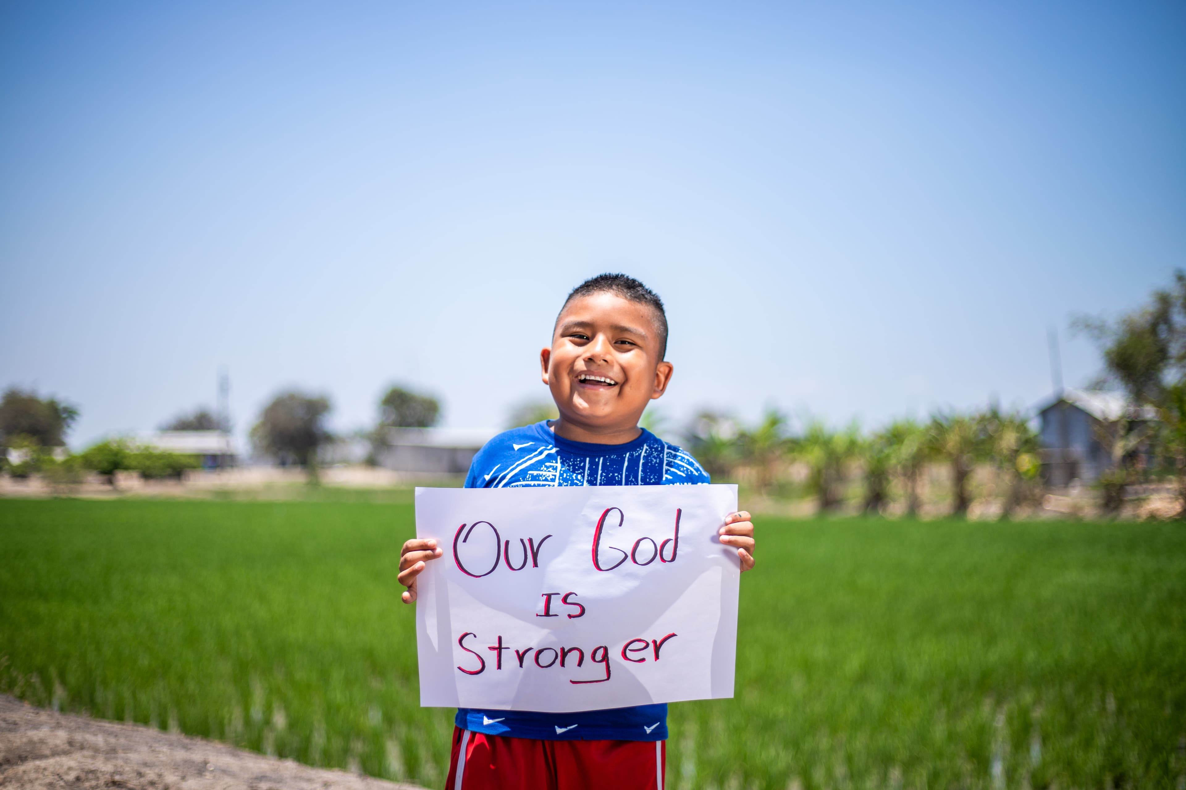 A young boy smiles while holding a sign in front of a green field.