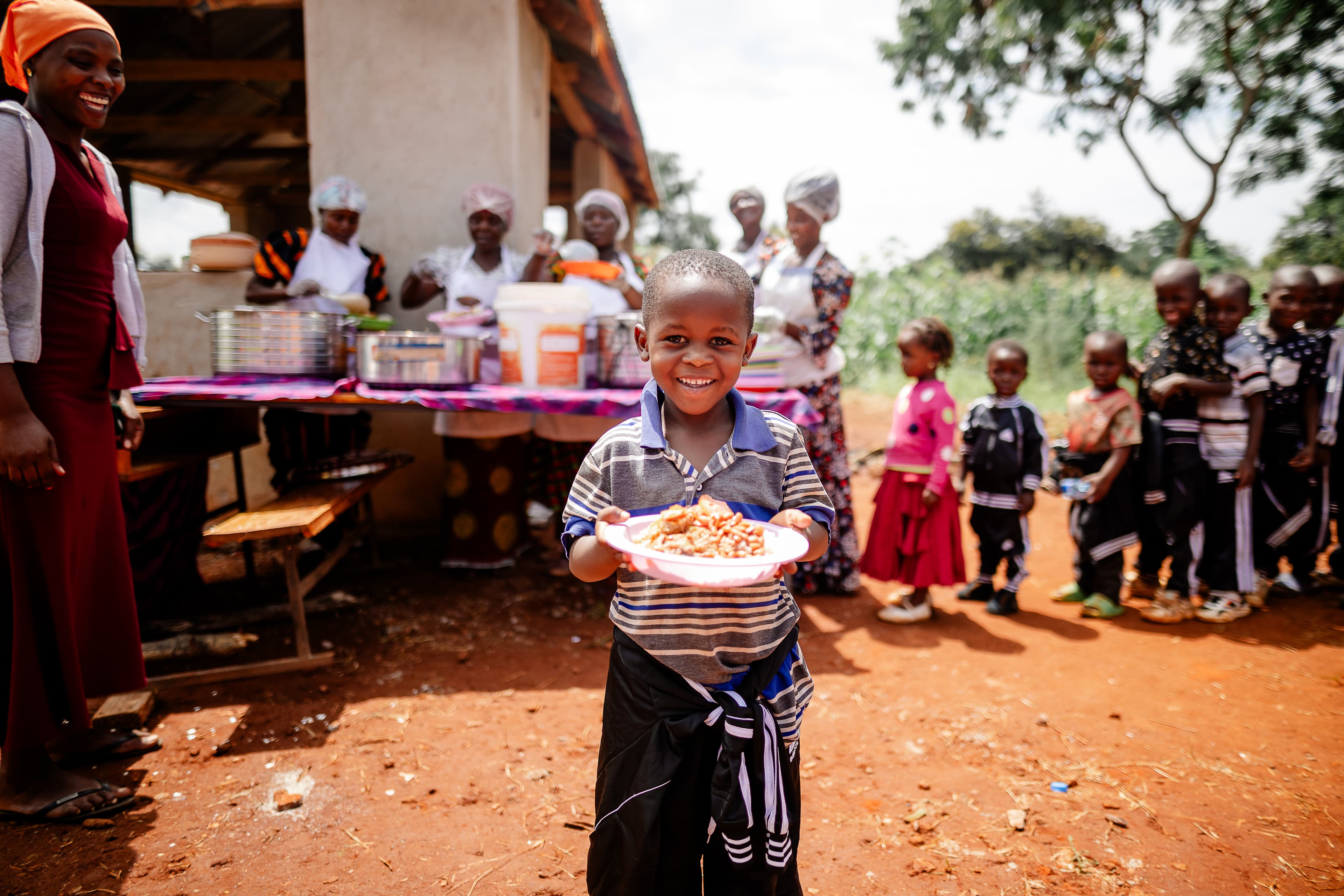 A young boy stands outside carrying a plate of food, with more children waiting in line for food behind him.
