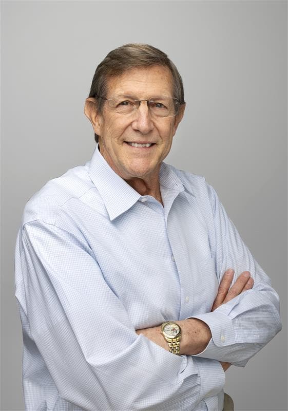 Wess Stafford in a blue shirt, wearing a gold watch, is smiling with his arms folded across his chest, with a grey background behind him.