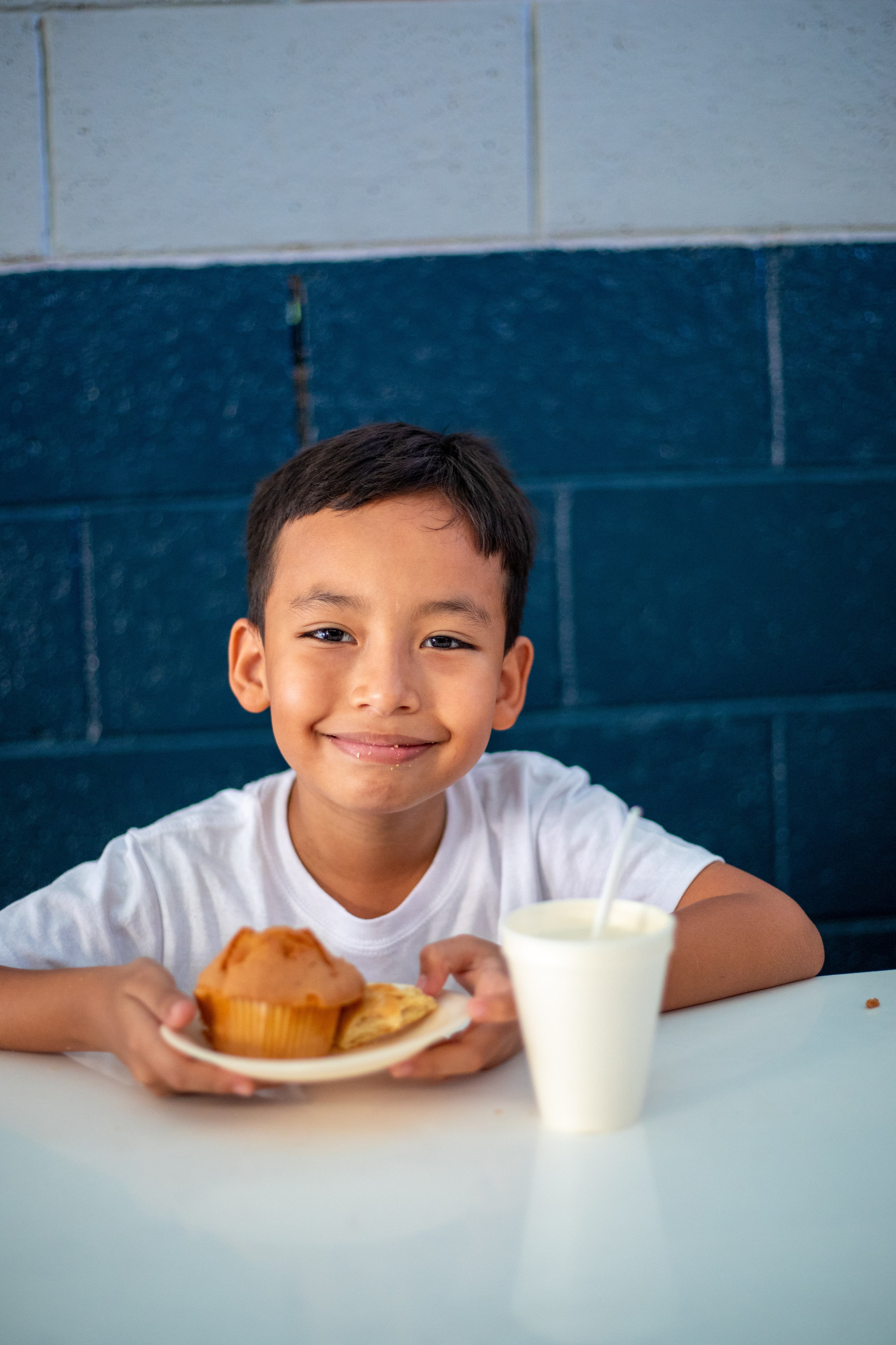 A boy sits at a white table at the center, wearing a white shirt, holding a small white plate of baked goods for a snack.