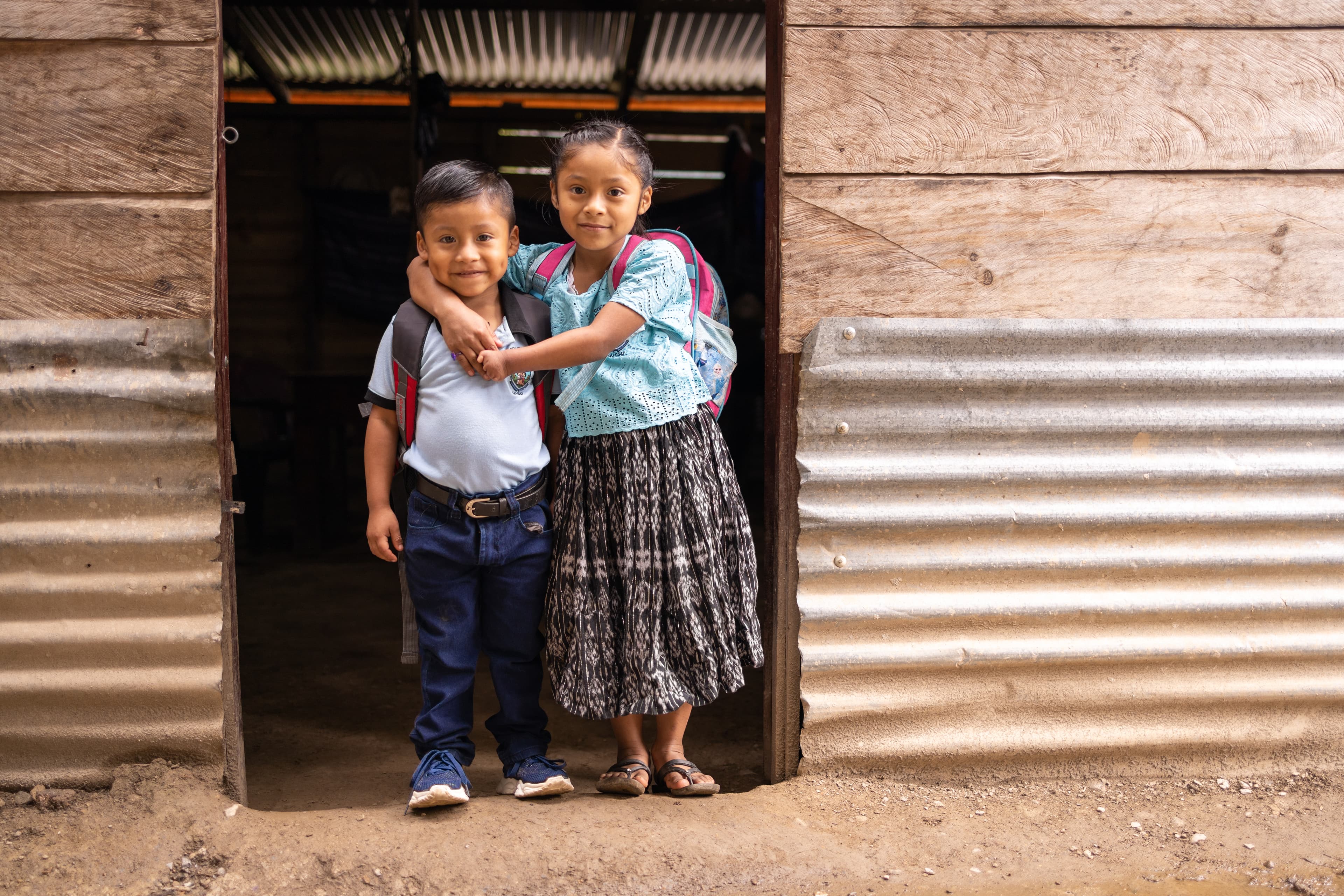 A young Guatemalan boy and girl stand in a doorway and embrace while smiling for the camera.