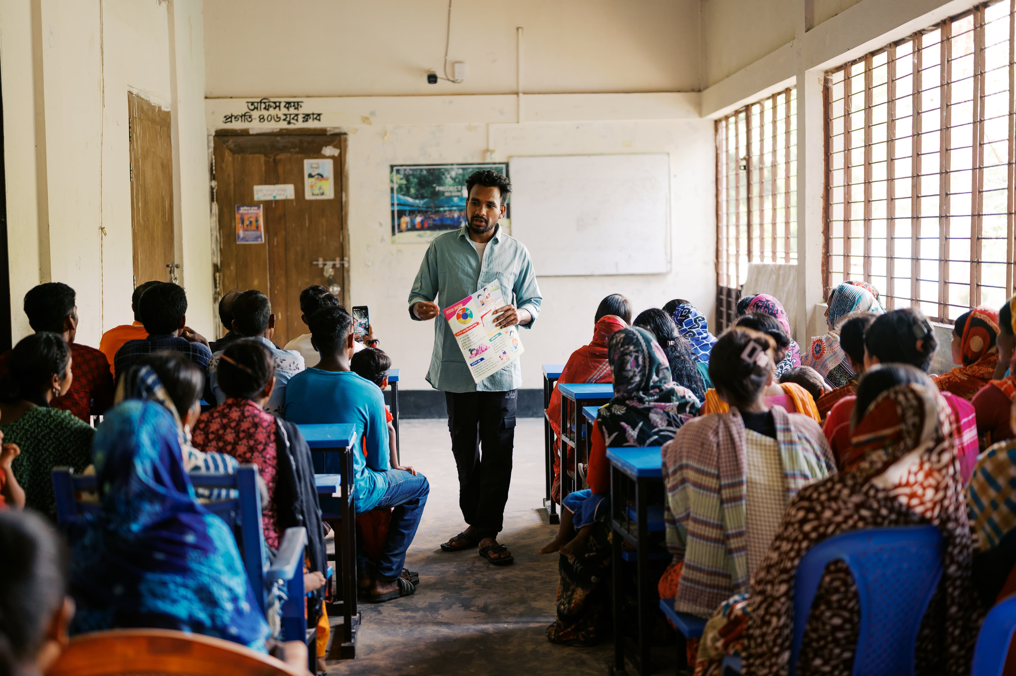 An adult male stands in front of a classroom of students while holding a poster.