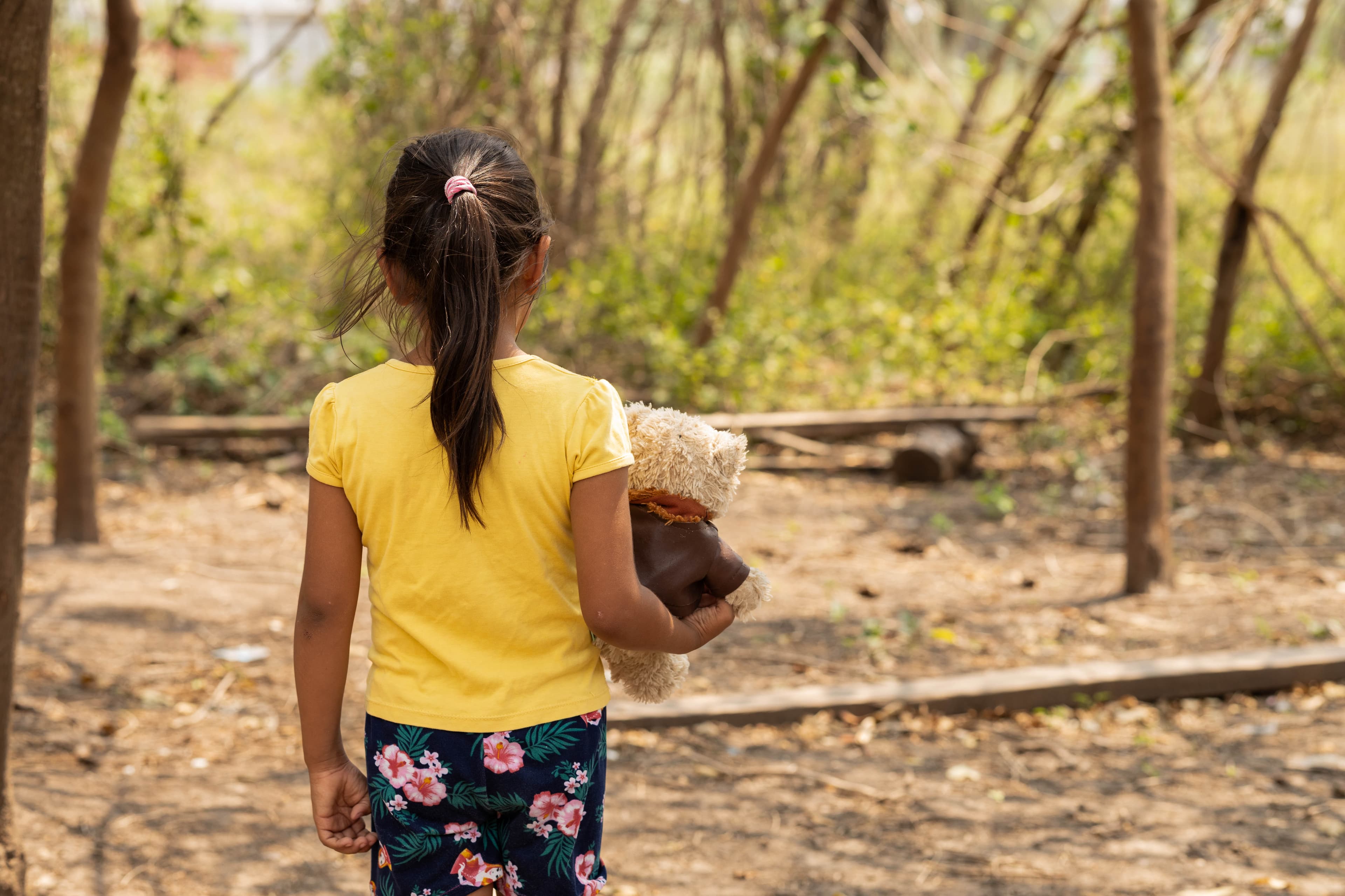 A young girl wearing a yellow shirt holds a teddy bear with her back turned to the camera.
