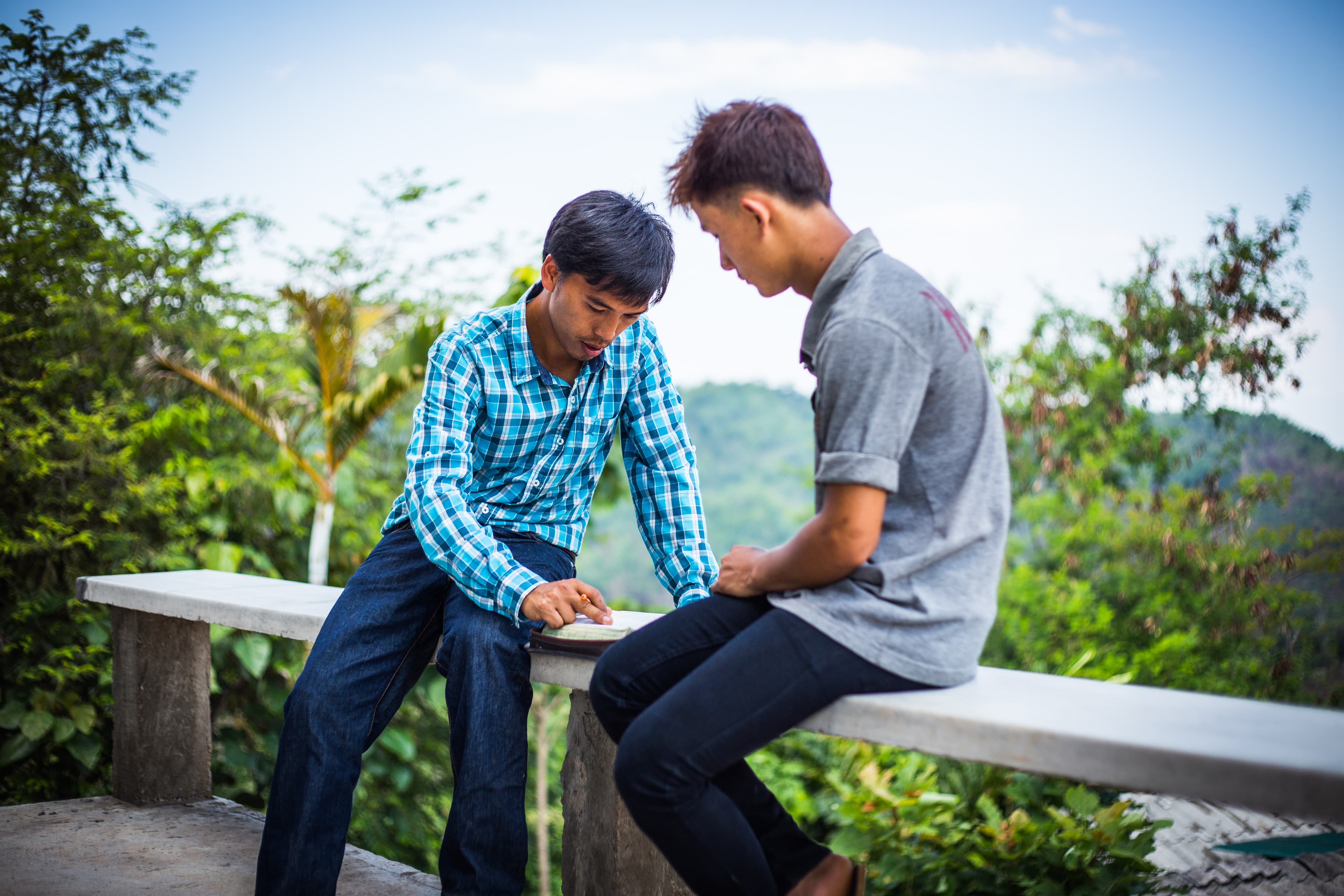 Two men sit on a bench leaning over to review and discuss scripture, surrounded by greenery.