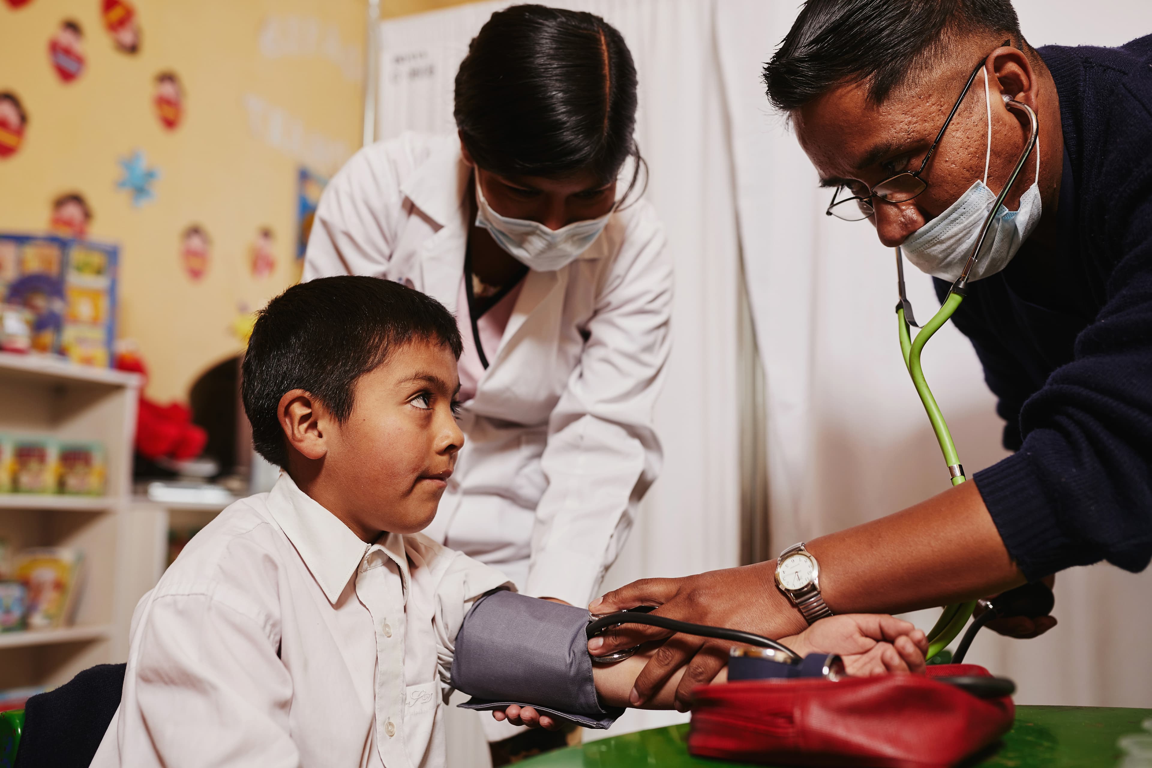Boy in white collared shirt sits, looks up at medical worker taking his blood pressure.