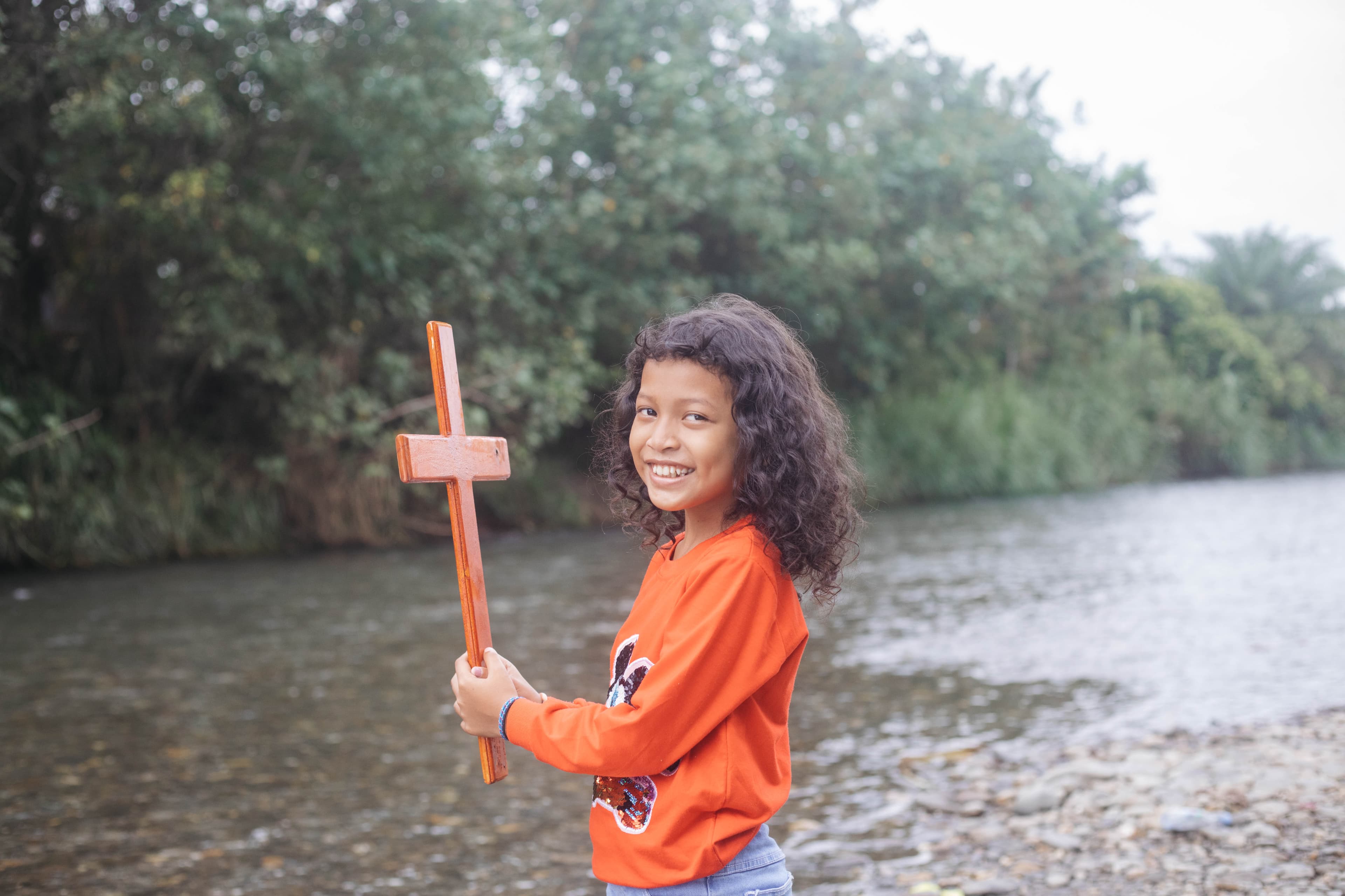 A young girl stands by a river holding a wooden cross and smiling for the camera.