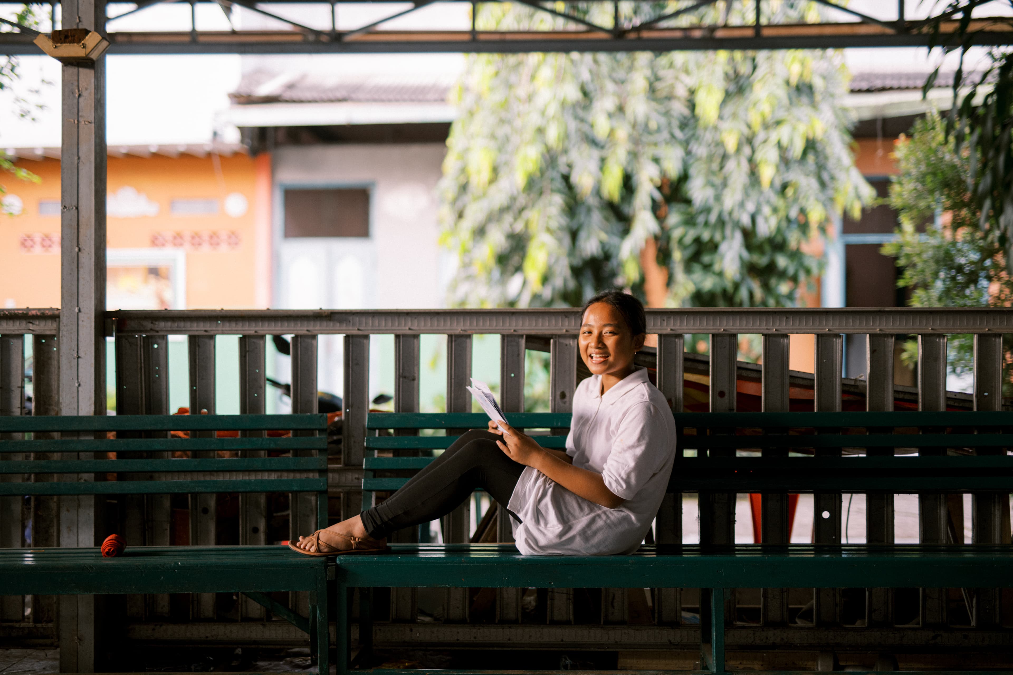 A young woman sits on a green bench and smiles at the camera holding letters from her sponsor.