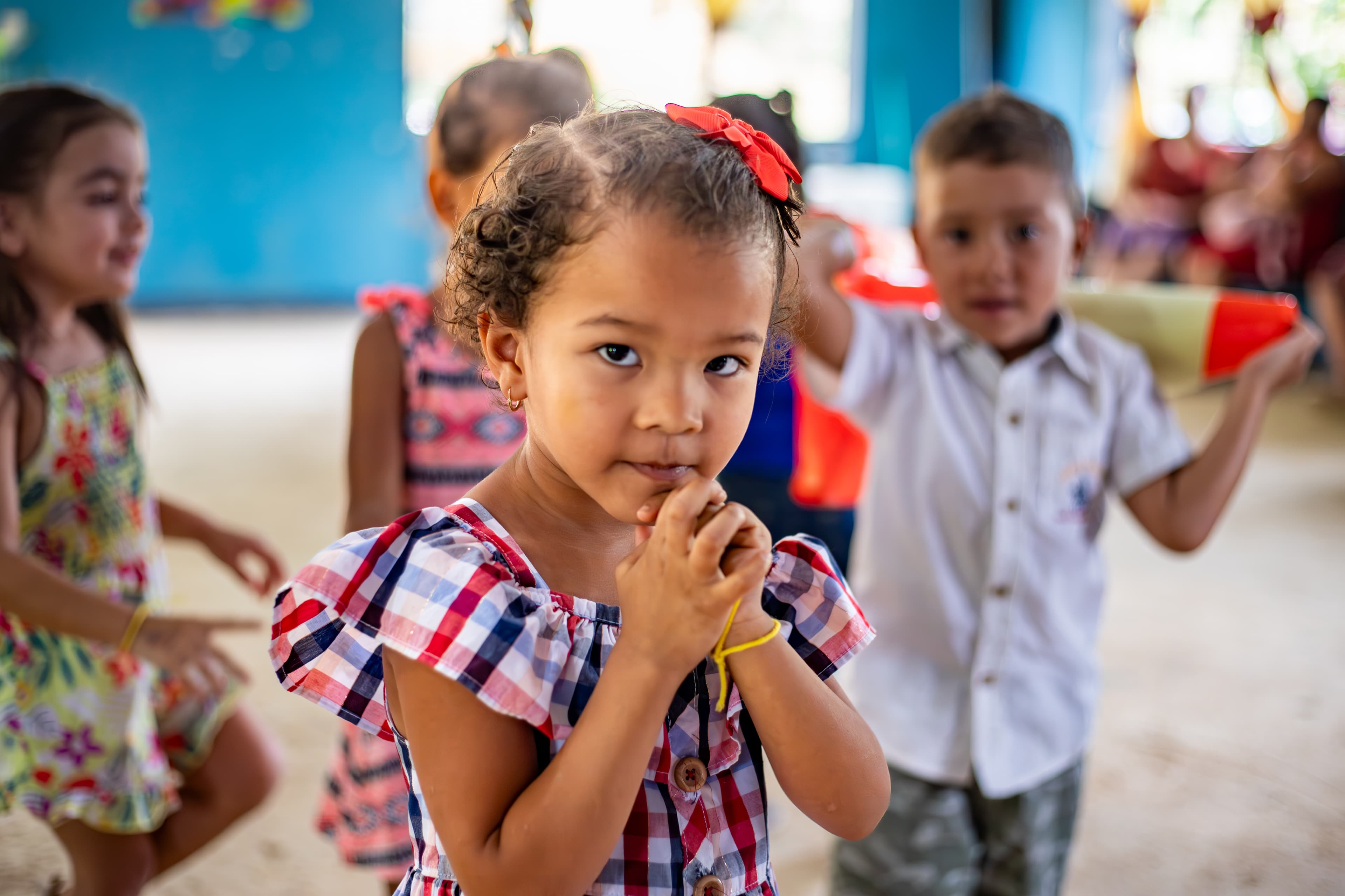 A young girl wearing a plaid dress looks at the camera with her hands clasped together. Other children are playing behind her.