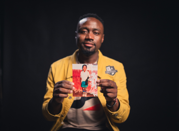 Kennedy, an alumni from Kenya, is wearing a yellow shirt and black pants. He is holding out a picture of himself as a child.
