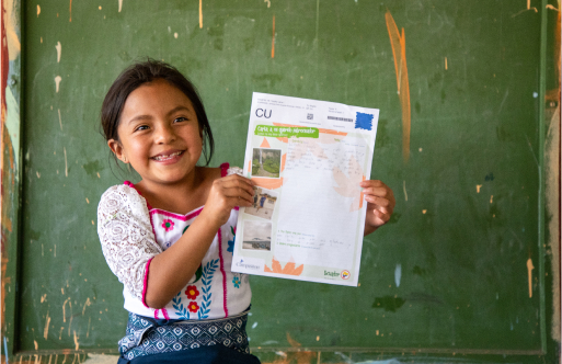 Lizbeth is wearing a white shirt with a floral pattern on it. She is standing in front of a green board in her classroom and she is holding up one of her sponsor's letters.